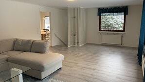 En moderne stue med en beige sofa, et glasbord og trægulv. Der er et vindue med udsigt til sneklædte træer.