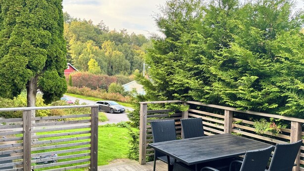 En træterrasse med et bord og stole, omgivet af frodig vegetation og udsigt til en vej og fjerne bygninger.