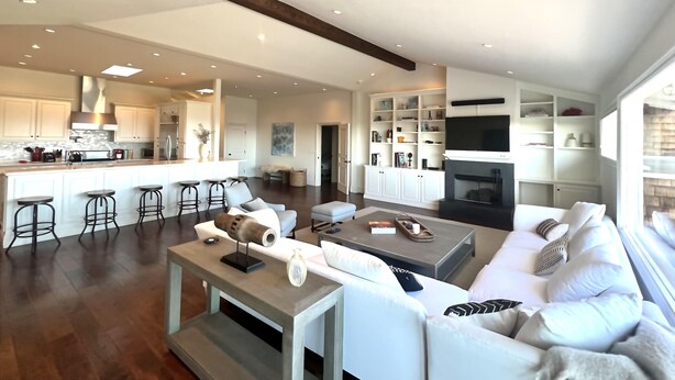 A modern living room with a sofa, coffee table, and bar stools. There is a kitchen area with a stove and cabinets.