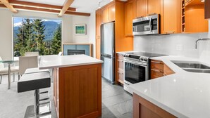 A modern kitchen with wooden cabinets, stainless steel appliances, and a view of mountains through a large window. - Newly Renovated Ski/Out Condo w/ Pool, Hot Tub & Steam Shower (Whistler)
