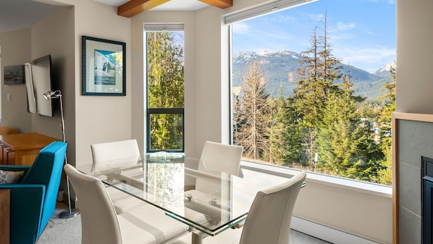 A modern room with a glass table and chairs, a TV, and a large window with a mountain view. - Newly Renovated Ski/Out Condo w/ Pool, Hot Tub & Steam Shower (Whistler)