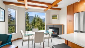 A modern kitchen with a fireplace, large windows offering a mountain view, and a dining area with a glass table. - Newly Renovated Ski/Out Condo w/ Pool, Hot Tub & Steam Shower (Whistler)