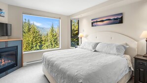 A bedroom with a large window, a fireplace, a bed with a white headboard, and a painting on the wall. - Newly Renovated Ski/Out Condo w/ Pool, Hot Tub & Steam Shower (Whistler)