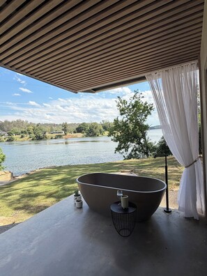 Ein modernes Badezimmer mit einer freistehenden Badewanne, einem kleinen Tisch mit Kerze und Blick auf einen See und Bäume.
