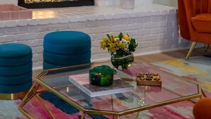 A living room with a white brick fireplace, blue ottomans, a glass coffee table, and a colorful rug.