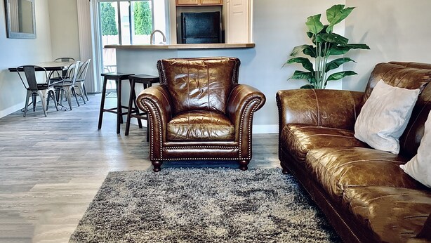 A living room with a brown leather armchair, a brown leather sofa, a dining table with chairs, a large plant, and a kitchen area with wooden cabinets and a TV. -  Spacious and Cozy with bonus room! (meridian)