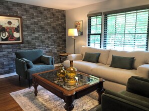 Ein Wohnzimmer mit Backsteinwand, blauem Sessel, beiger Couch, einem Glastisch mit dekorativen Vasen und einem Fenster mit Jalousien.