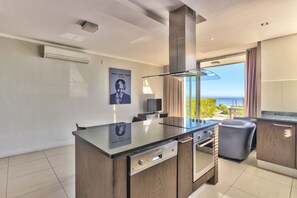 A modern kitchen with a central island, built-in oven, and a large window offering a view of the sea.