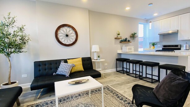 A modern living room with a black sofa, a coffee table, and a patterned rug. There is a dining area with bar stools and a kitchen with white cabinets and appliances.