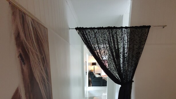 A hallway with a large framed picture on the left, a black lace curtain on the right, and a glimpse of a room with a chair and lamp in the distance. - Farm Stay Cottage in the beautiful Gold coast hinterland (Illinbah)