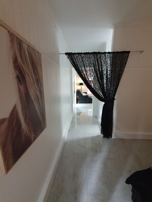 A hallway with a large framed picture on the left, a black lace curtain on the right, and a glimpse of a room with a chair and lamp in the distance. - Farm Stay Cottage in the beautiful Gold coast hinterland (Illinbah)