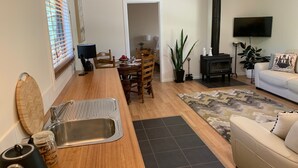 Een moderne woonkamer met een bank, een eethoek met houten stoelen, een keuken met een spoelbak en waterkoker, een open haard en een aan de muur bevestigde televisie.