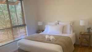Een keurig opgemaakt bed met een beige sprei, twee opgerolde handdoeken en witte kussens. Er staan twee nachtkastjes met lampen en een klein decoratief object.