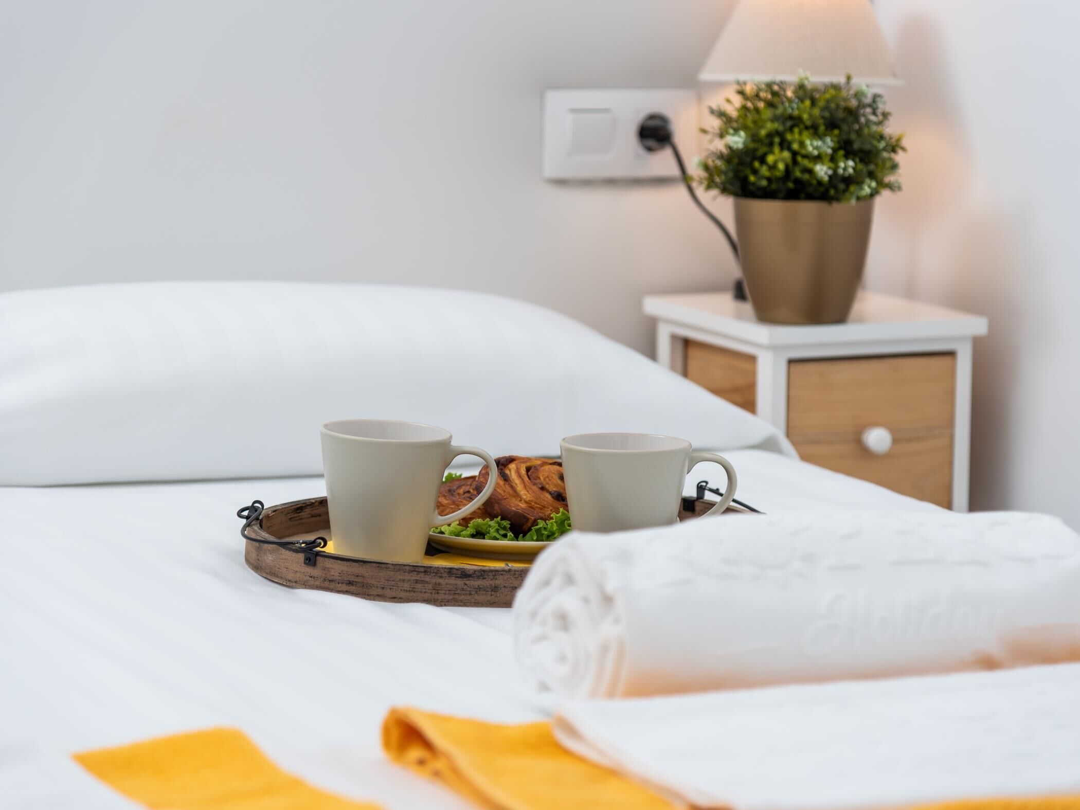 A neatly made bed with a white pillow, a tray with two mugs and a pastry, a small bedside table with a lamp and a plant, and a yellow towel.