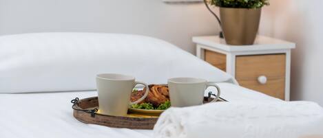 A neatly made bed with a white pillow, a tray with two mugs and a pastry, a small bedside table with a lamp and a plant, and a yellow towel.