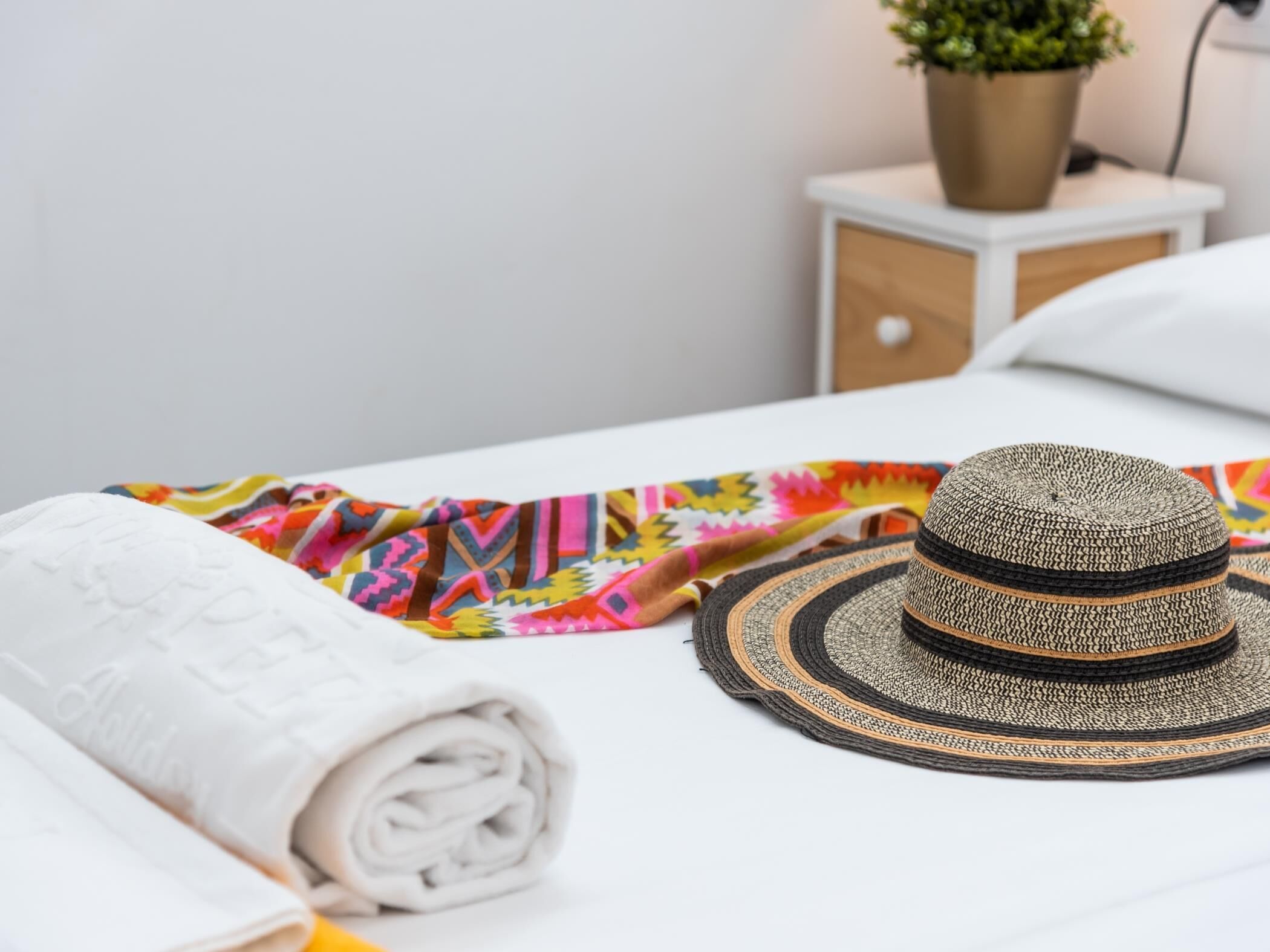 A neatly made bed with a rolled white towel, a colorful patterned cloth, and a woven hat on a white nightstand with a potted plant.