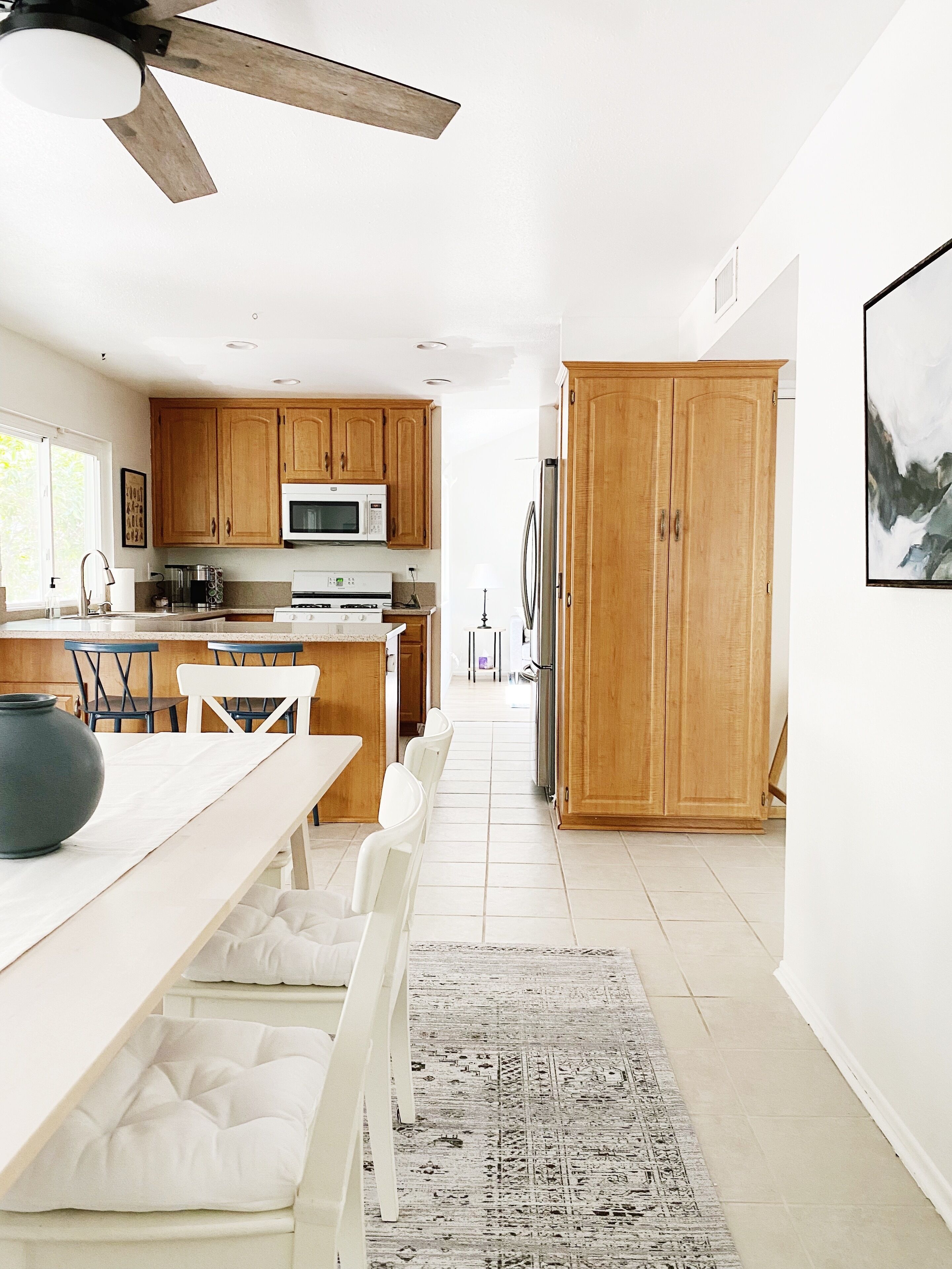A modern kitchen with wooden cabinets, a dining area with a white table and chairs, and a large rug leading to a door.