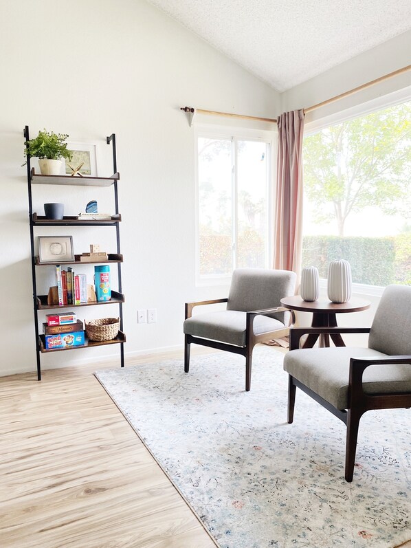 Una habitación con piso de madera, una estantería, dos sillas, una mesa y un ventanal con cortinas.