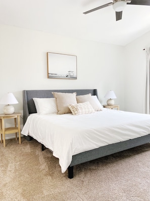 Un dormitorio ordenado con una cama grande, dos mesitas de noche con lámparas, un ventilador de techo y un cuadro enmarcado en la pared.