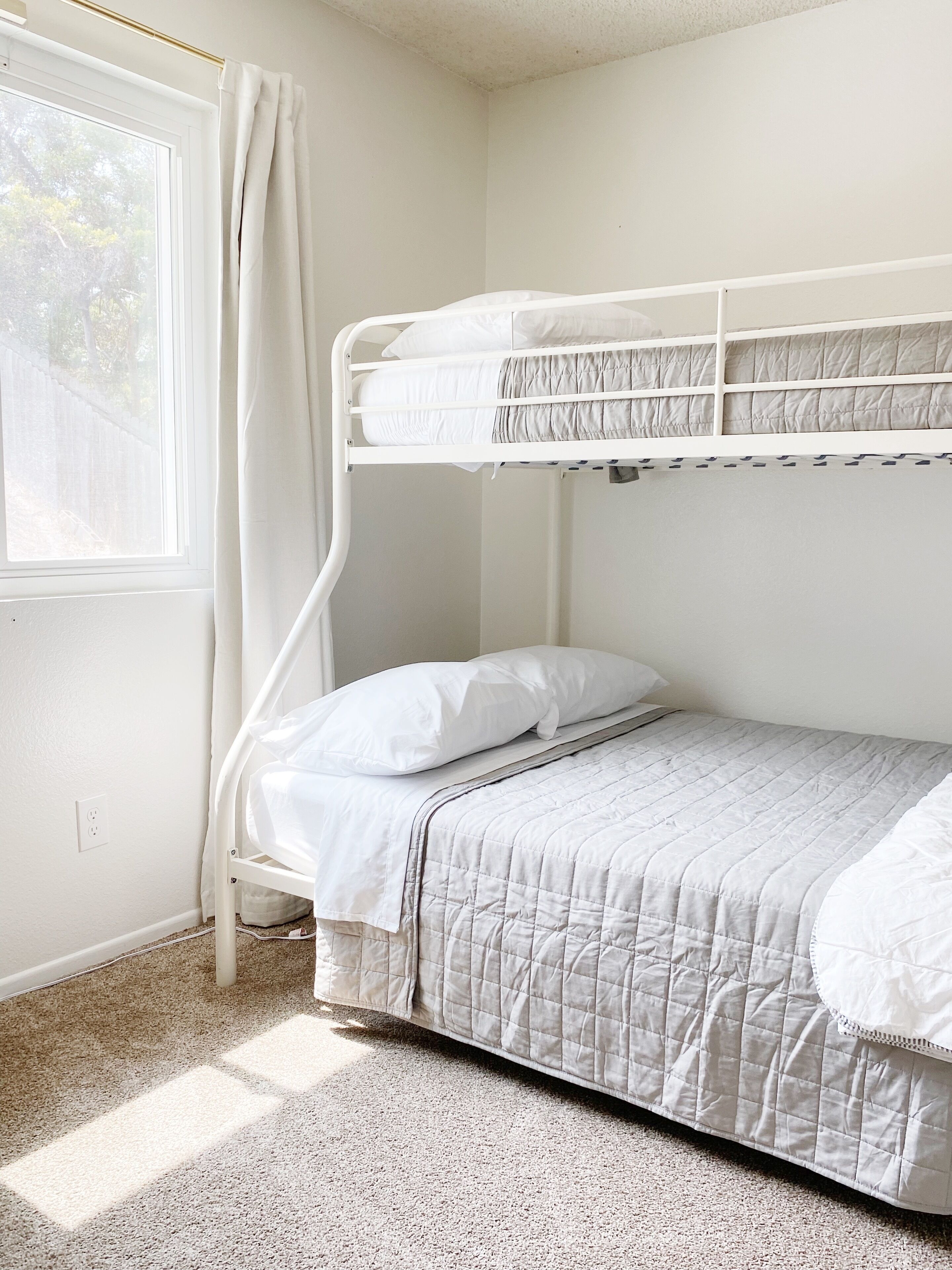 A bunk bed with a quilted bedspread and white bedding in a room with a window and a curtain.