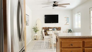 A kitchen with a stainless steel refrigerator, wooden cabinets, a ceiling fan, a dining area with a surfboard, and a TV mounted on the wall. - The perfect stay for Families and Friends (Oceanside)