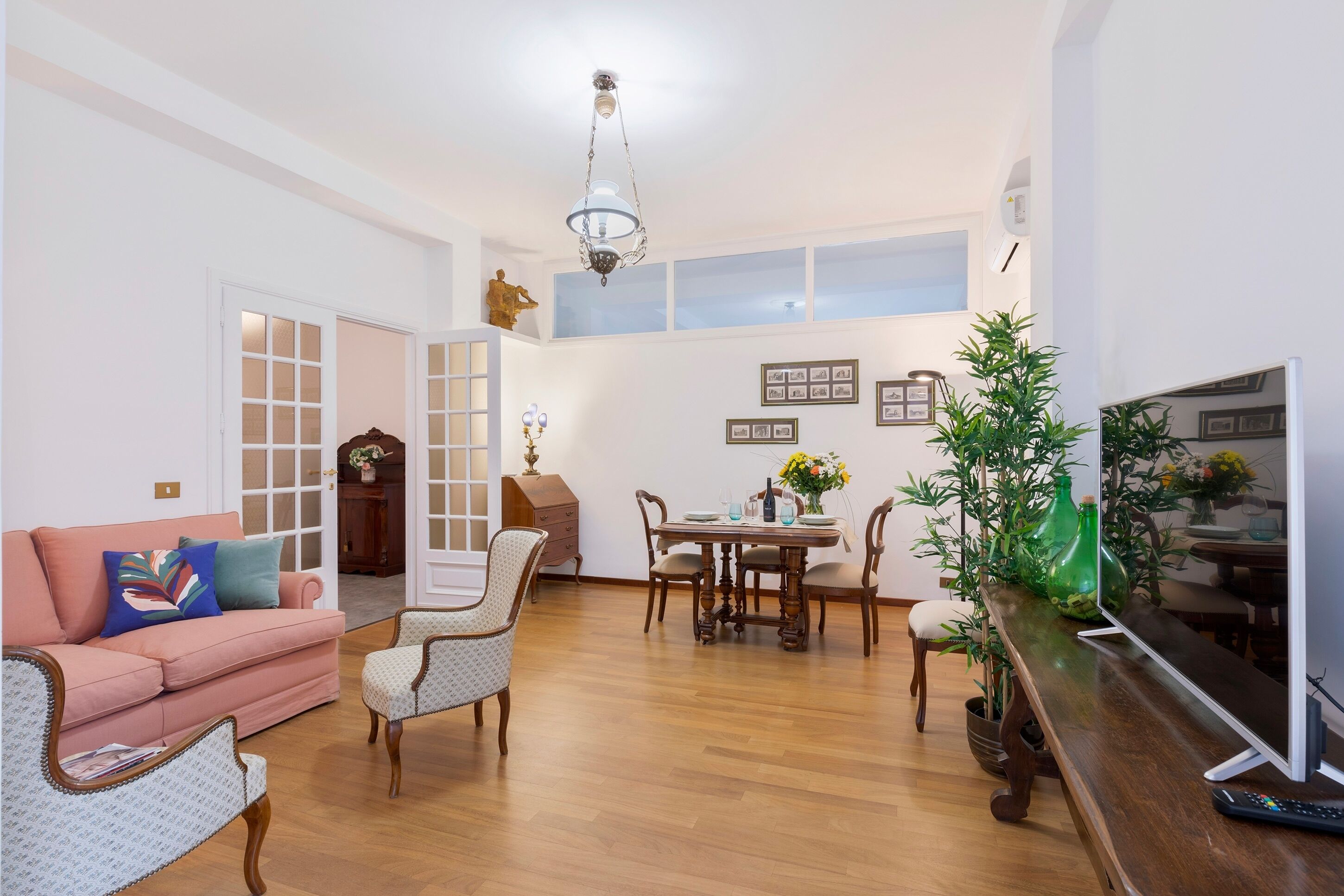 Una sala de estar con un sofá rosa, sillas estampadas, una mesa de comedor de madera con sillas, un televisor y una planta.