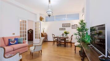 Una sala de estar con un sofá rosa, sillas estampadas, una mesa de comedor de madera con sillas, un televisor y una planta.