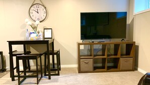 Un salon avec un meuble TV en bois, un grand téléviseur à écran plat, une horloge murale et une petite table avec une chaise.