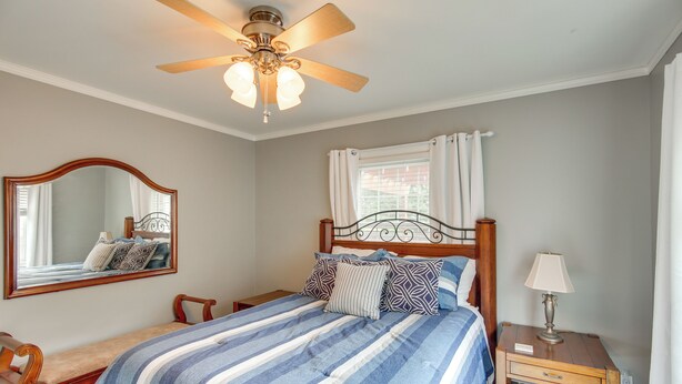 Un dormitorio con cama, mesita de noche, lámpara, ventilador de techo, espejo y ventana con cortinas.