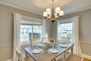 Une salle à manger avec une table en bois dressée pour quatre personnes, un lustre et de grandes fenêtres ornées de rideaux blancs.