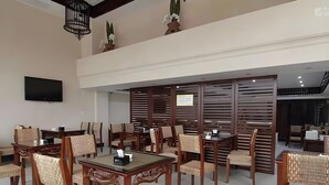 A dining area with wooden tables and chairs, a flat-screen TV on the wall, and decorative plants.