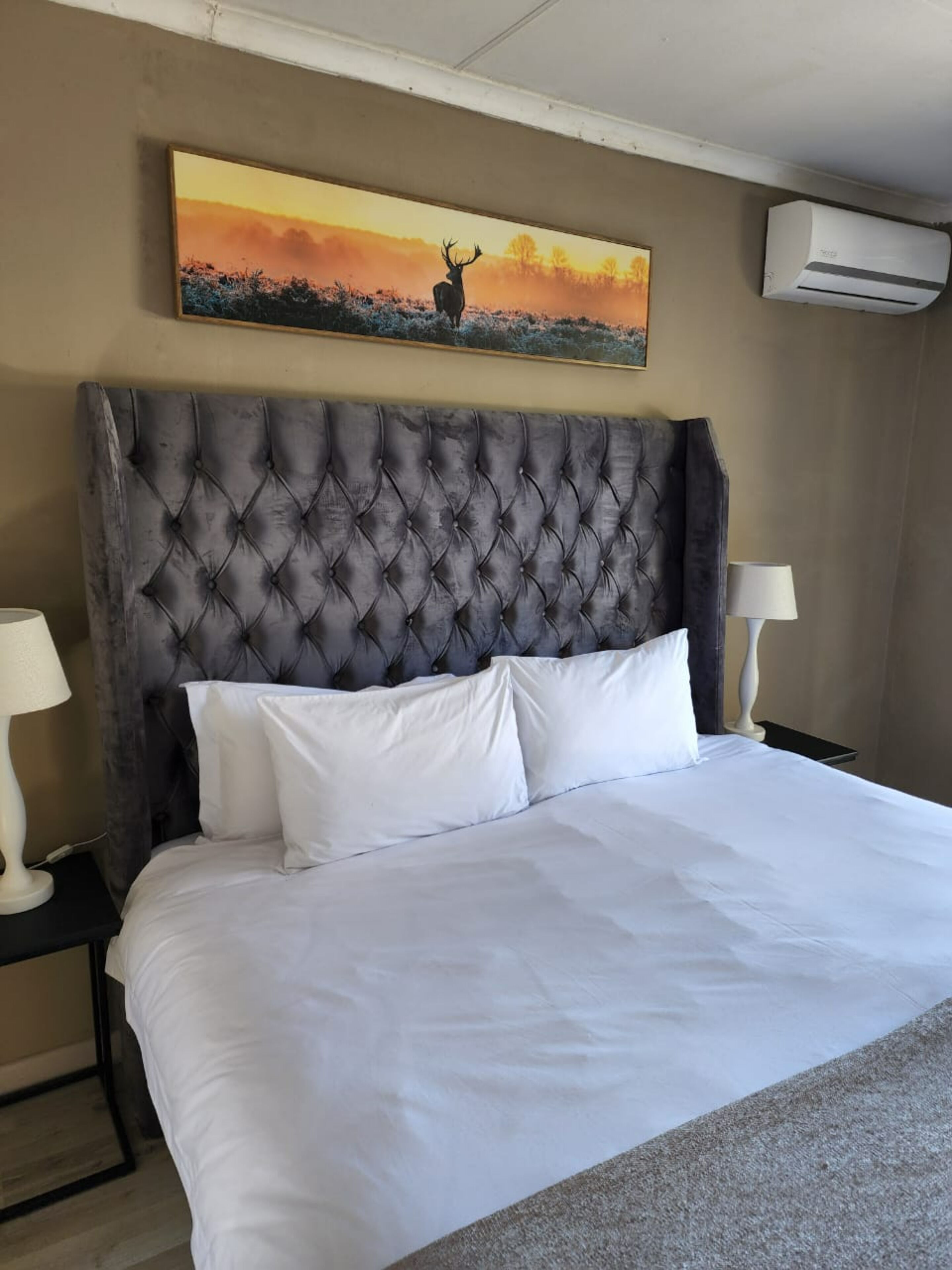 A hotel room with a large bed, a tufted headboard, white bedding, two bedside lamps, and a painting of a deer by a sunset.