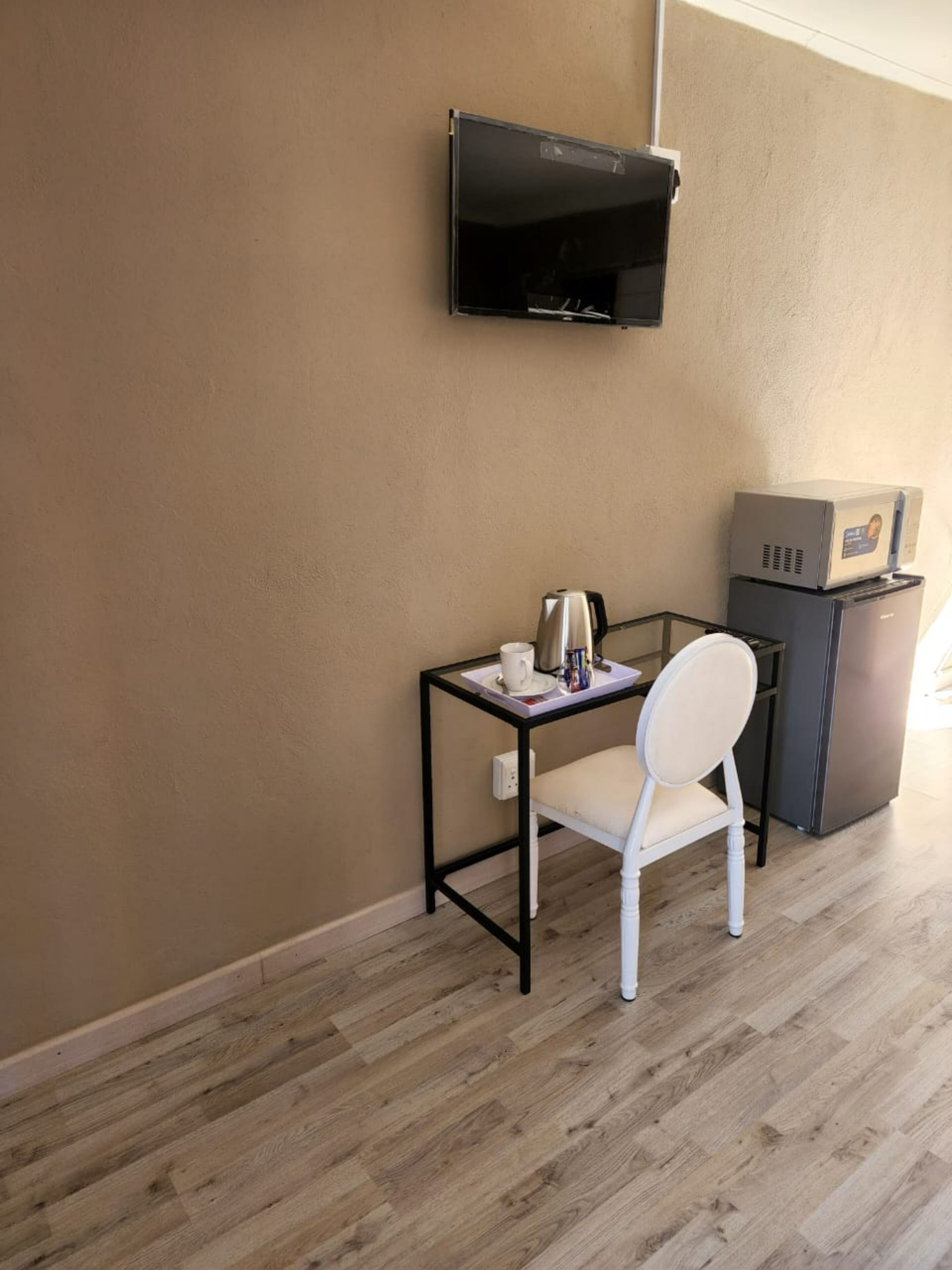 A small room with a wooden floor, a white chair, a small table with a cup and a kettle, a television mounted on the wall, and a refrigerator with a box on top.