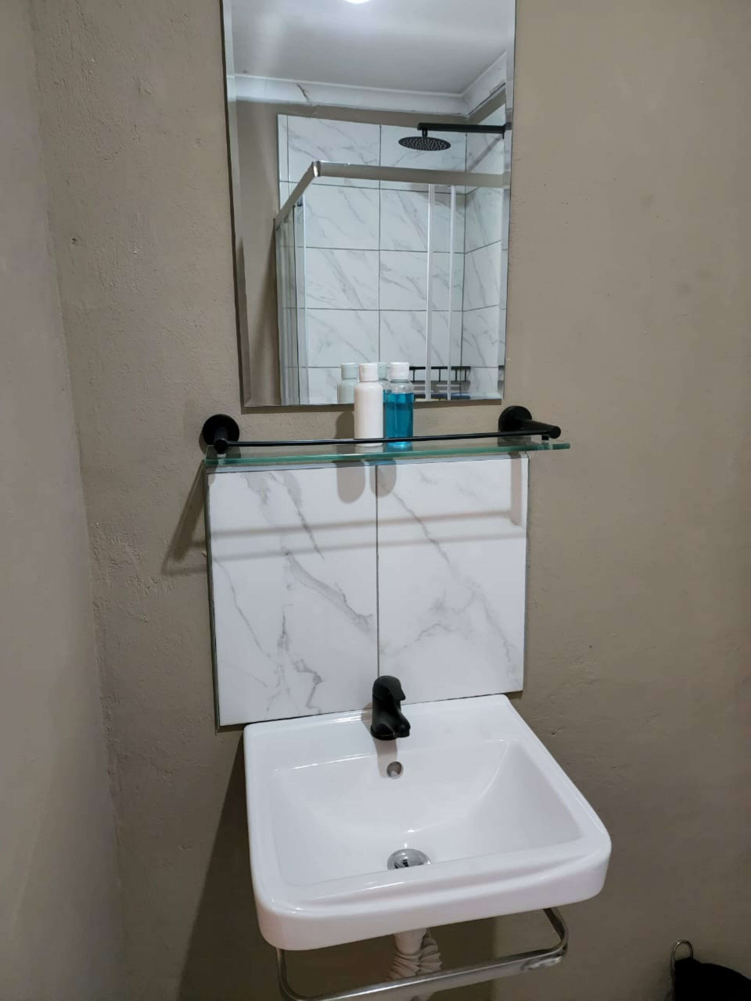 A bathroom with a white sink, a glass shelf with toiletries, and a mirror reflecting a shower area.