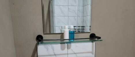 A bathroom with a white sink, a glass shelf with toiletries, and a mirror reflecting a shower area.