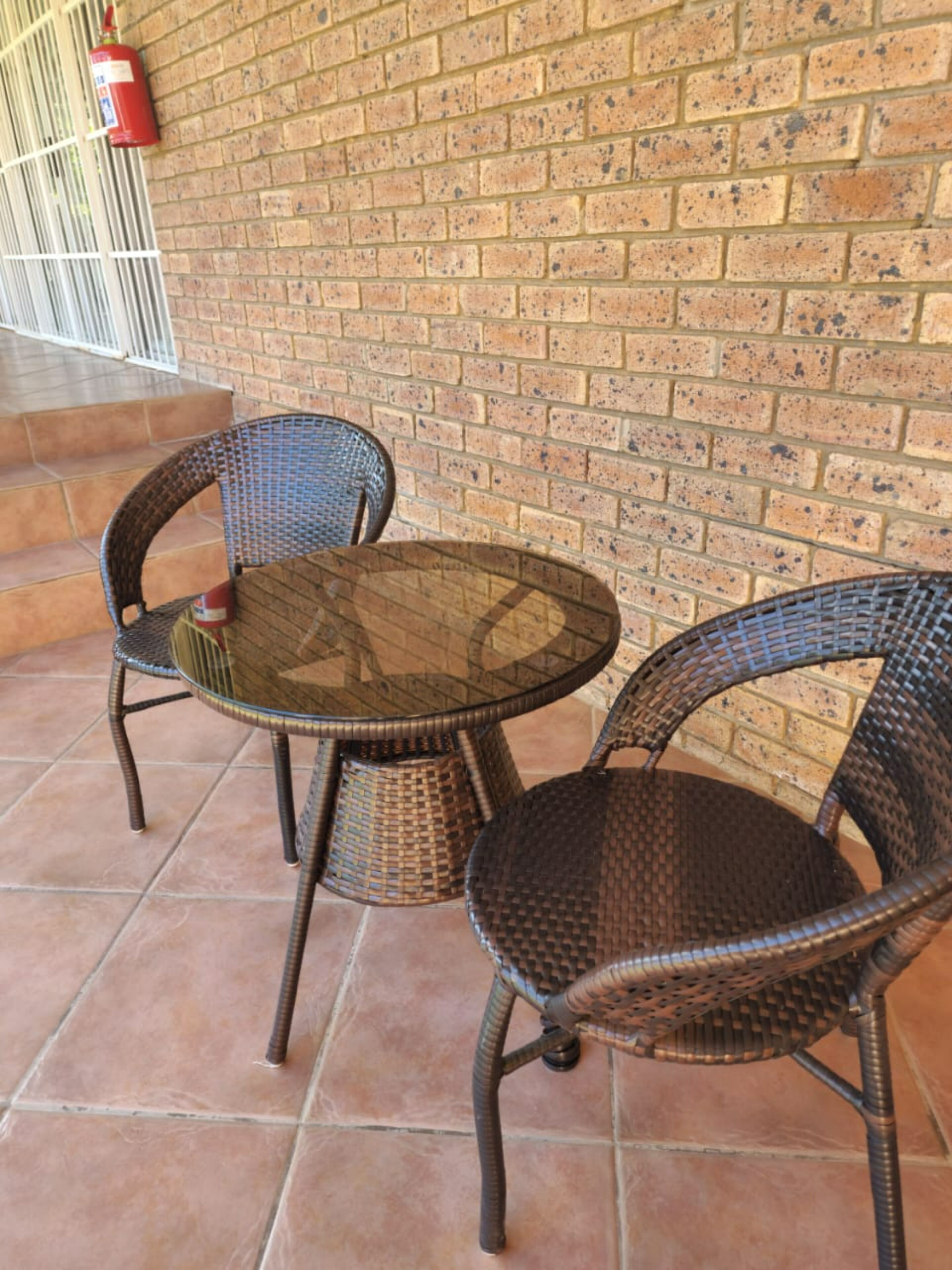 A patio area with two wicker chairs and a glass table, set against a brick wall.