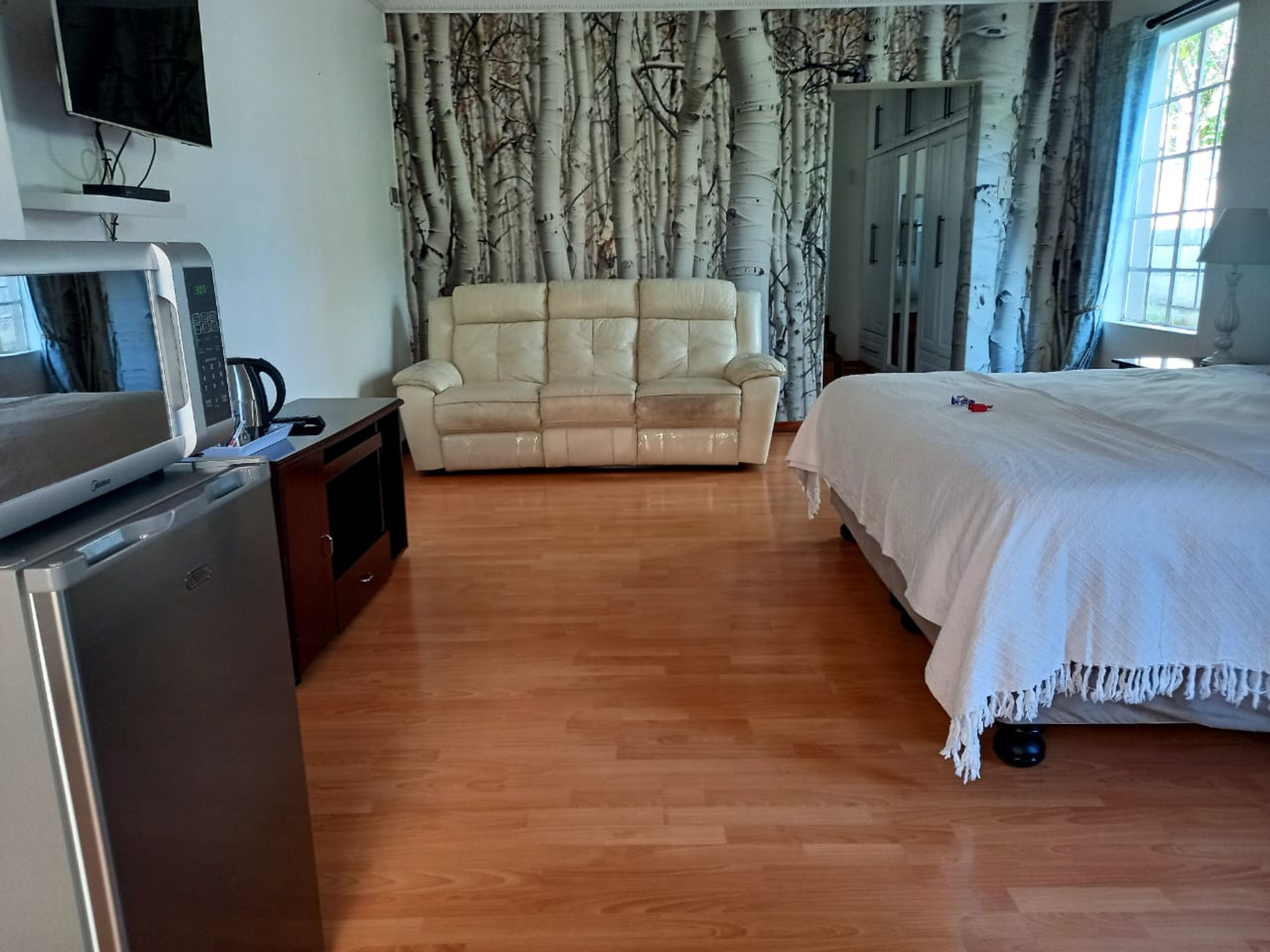 A hotel room with a wooden floor, a beige sofa, a bed with white bedding, a TV on a stand, and a wardrobe.