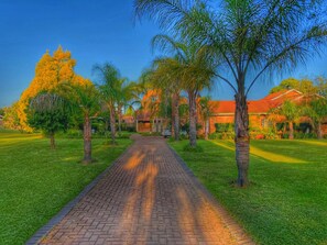 A paved pathway leading to a building with a red roof, surrounded by palm trees and well-maintained grass. - The Hills Guest House (Newcastle)