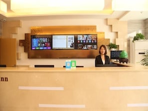 A reception desk with a digital display board above it, a receptionist, and a small plant on the counter.