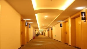 Un couloir d'hôtel avec un sol recouvert de moquette, des portes en bois et des luminaires encastrés au plafond.