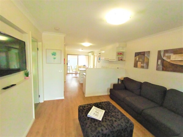 A living room with a sofa, a television, and a small table with a book. There is a kitchen area in the background with a countertop and cabinets. - My Abode in Albury * Close to Charles Sturt University & Thurgoona Golf Club (Thurgoona)