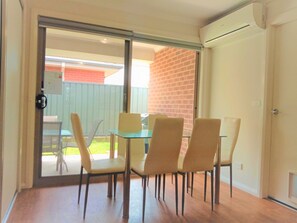 A dining area with a glass table and four chairs, a sliding glass door leading to an outdoor patio, and a brick wall.