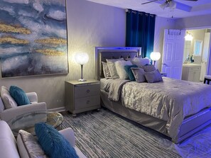 A modern bedroom with a large bed, a nightstand with a lamp, a glass coffee table, and a painting on the wall.