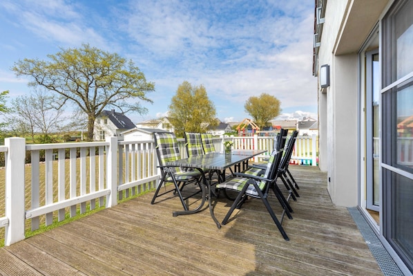 A wooden deck with a table and chairs, a white fence, and a building with large windows.
