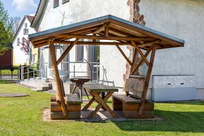 Ein Holz-Sitzbereich im Freien mit Tisch und Bänken, eine Steinmauer und ein Gebäude im Hintergrund.