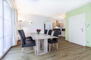 A dining area with a table and chairs, a vase with flowers, a kitchen in the background, and a door with a light switch.