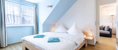 A bedroom with a sloped ceiling, a bed with white linens, a bedside table with a lamp, and a view of a living area.