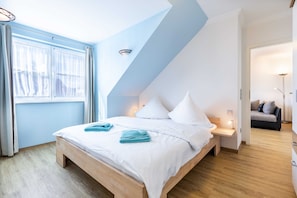A bedroom with a sloped ceiling, a bed with white linens, a bedside table with a lamp, and a view of a living area.