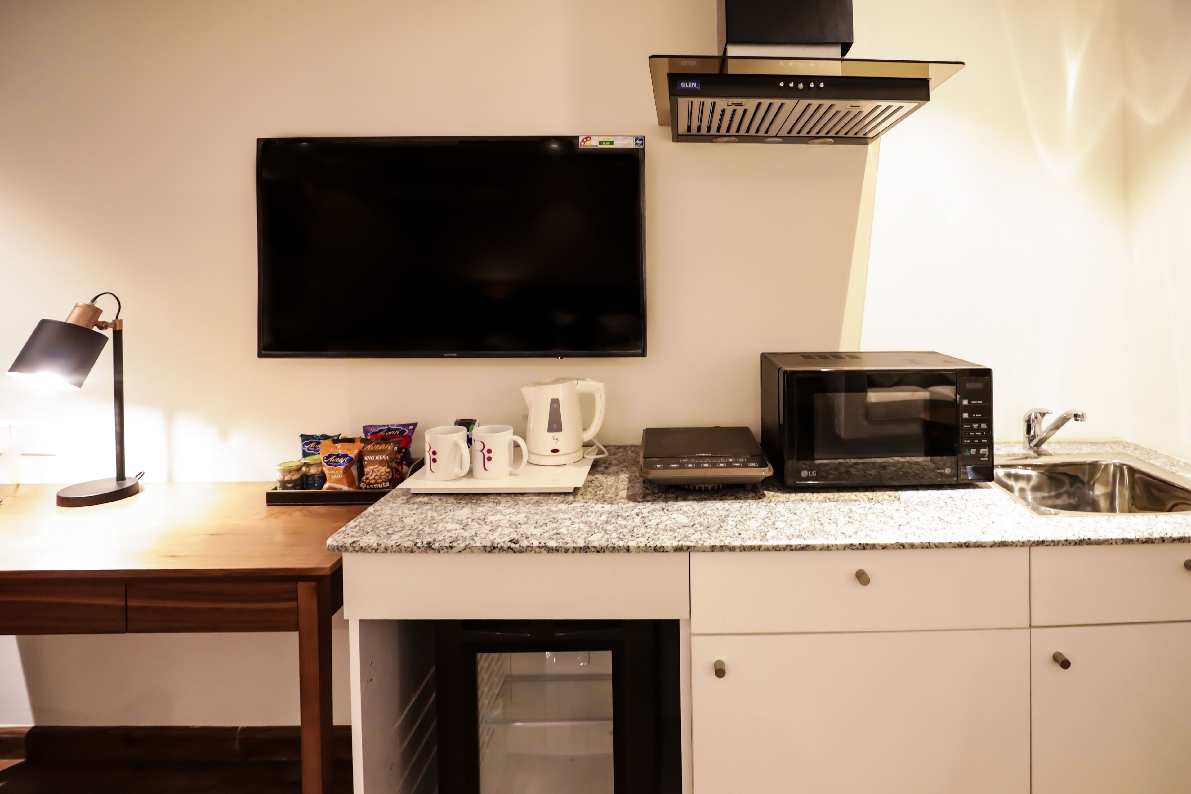 A kitchenette with a flat-screen TV, microwave, kettle, and coffee maker on a granite countertop.