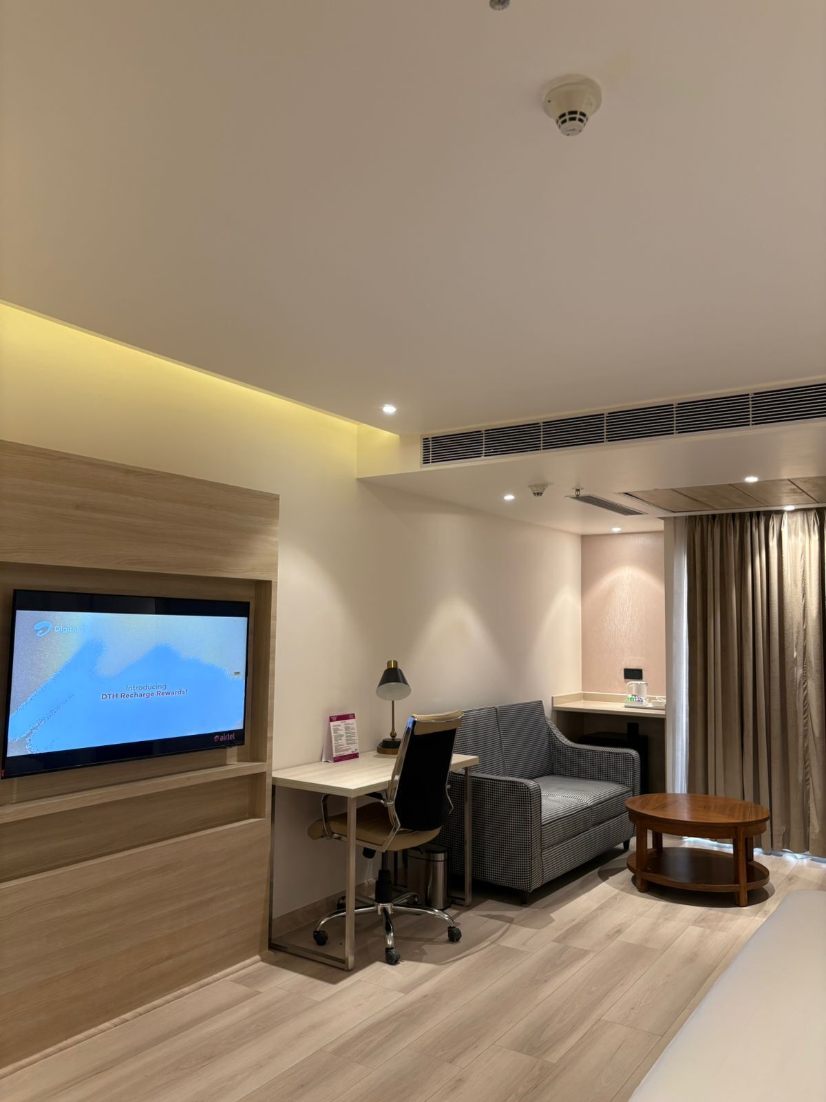 A modern hotel room with a flat-screen TV, a desk, a chair, a sofa, a small table, and a window with curtains.